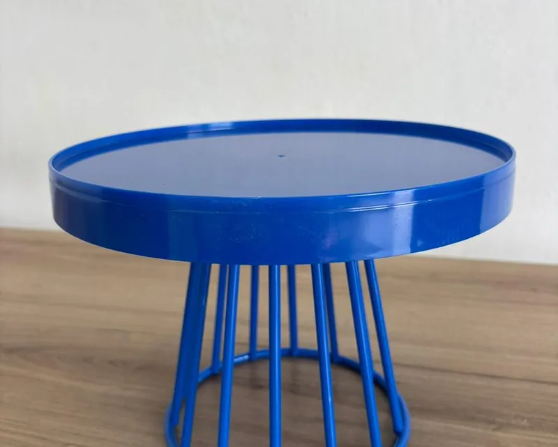 Boleira de metal azul royal, com tampo redondo de borda alta e base vazada de hastes, sobre uma bancada de madeira