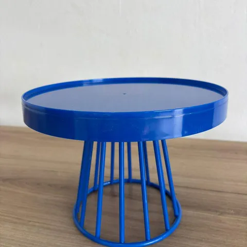 Boleira de metal azul royal, com tampo redondo de borda alta e base vazada de hastes, sobre uma bancada de madeira
