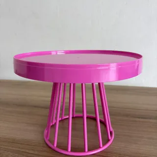 Boleira de metal pink, com tampo redondo de borda alta e base vazada de hastes, sobre uma bancada de madeira