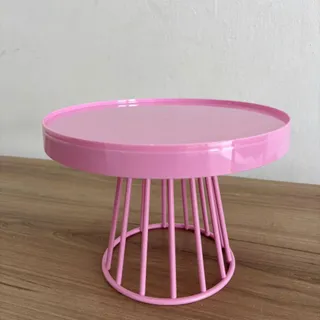 Boleira de metal rosa claro, com tampo redondo de borda alta e base vazada de hastes, sobre uma bancada de madeira