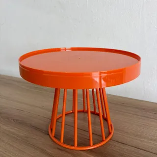 Boleira de metal laranja, com tampo redondo de borda alta e base vazada de hastes, sobre uma bancada de madeira
